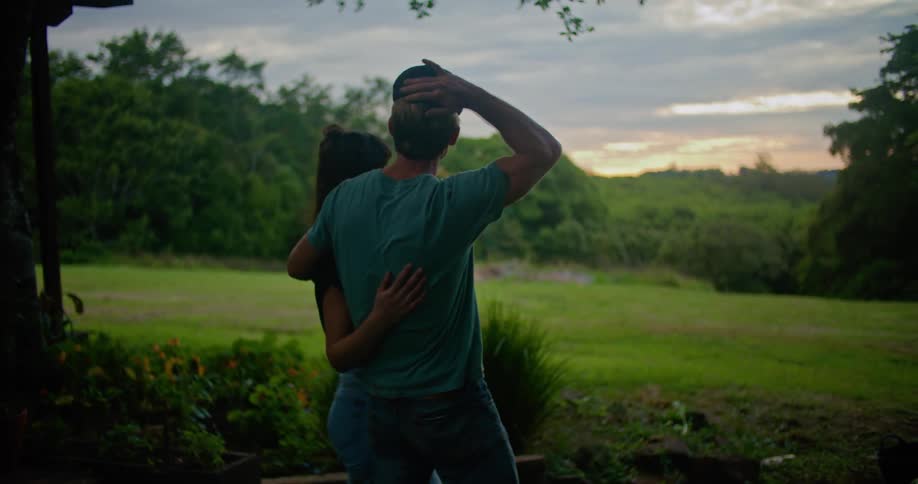 Man with dark blue cap turquoise t shirt and tattoo holds woman next to country house looking at green field in the countryside