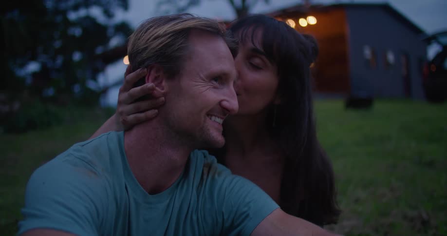 Woman with brown wavy hair and black blouse touches and kisses man with turquoise t shirt sitting on grass in frotn of country house at dusk
