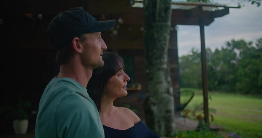 Man with dark blue cap turquoise t shirt and tattoo holds woman next to country house looking at green field in the countryside