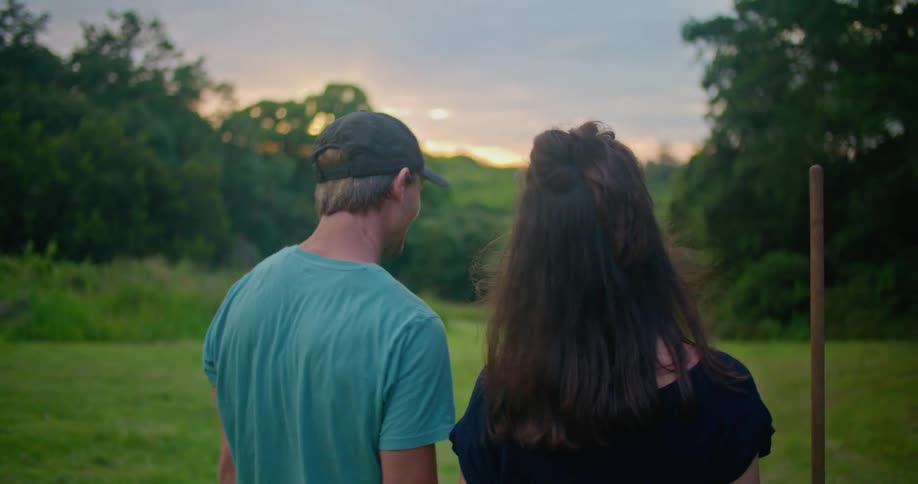 Man and woman carrying lawn tool walk across mowed field toward woods as sun shines past trees in red sky
