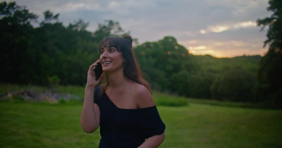 Woman with brown wavy hair and black dress talks on smartphone in the countryside next to forest at dusk