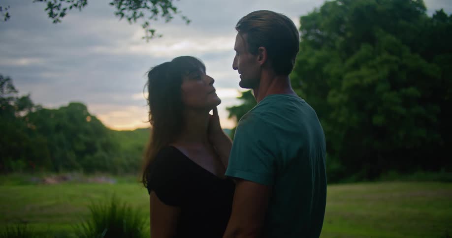 Man with turquoise t shirt holds woman with brown wavy hair and black dress lovingly looking into each other and making out in the countryside at dusk
