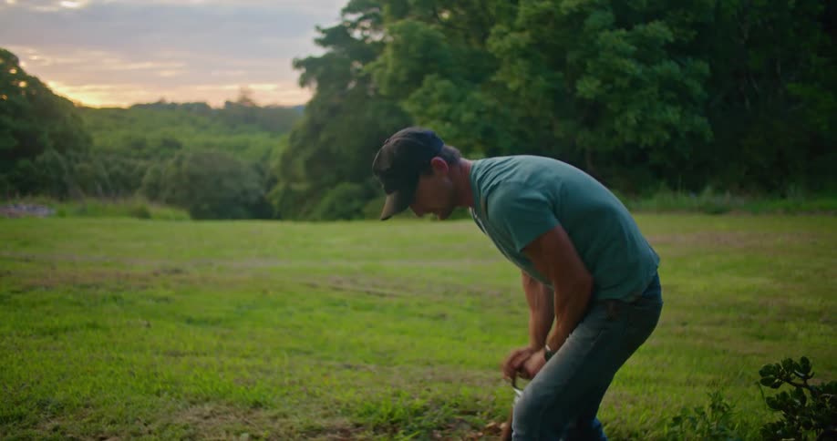 Man with dark blue cap turquoise t shirt jeans and tattoo digs in the garden using shovel in the countryside