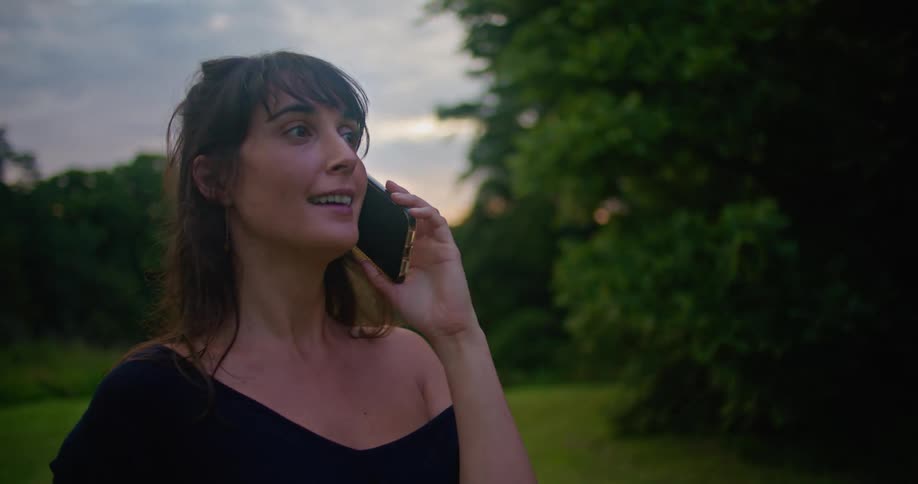 Woman with brown wavy hair and black dress talks on smartphone in the countryside next to trees at dusk