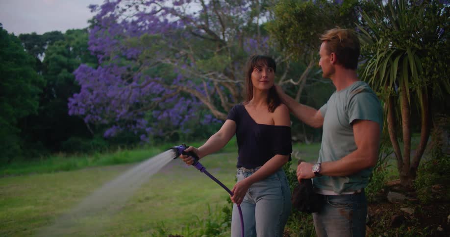 A woman using a hose to water a yard pauses to share a kiss with the man standing at her side