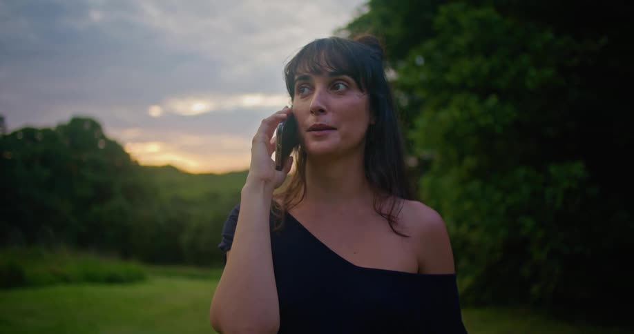A woman is standing outside on a lawn and talking on a cell phone at sunset