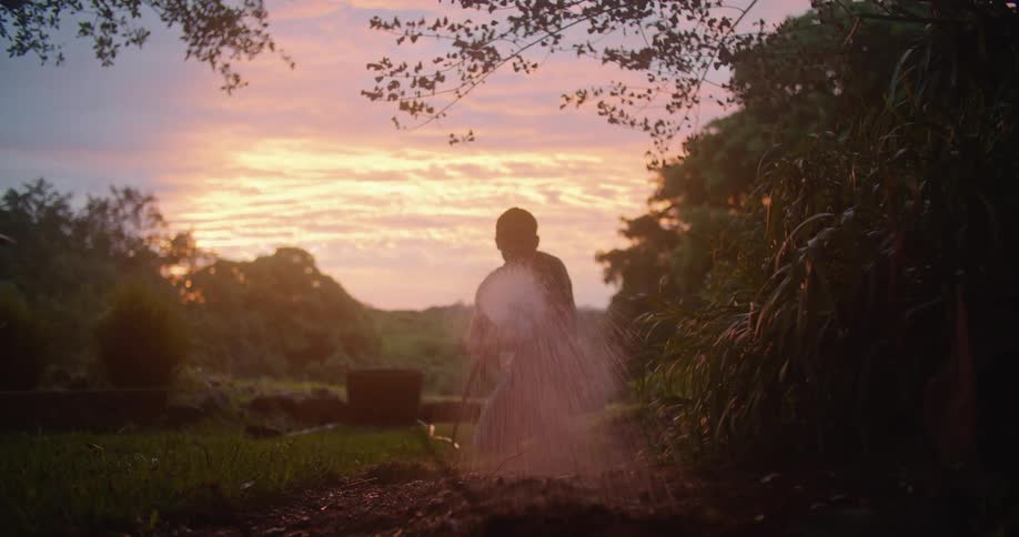 A man uses a hose to water soil while kneeling outdoors in the evening