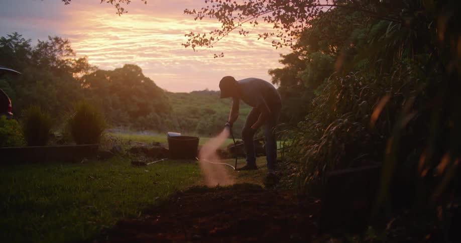Man waters ground with hose near parked car under a red sky