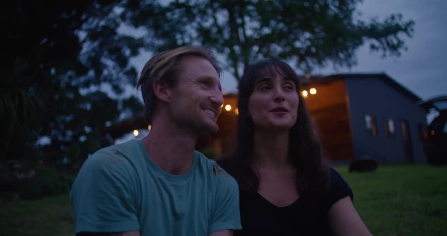 Man with turquoise t shirt and woman with brown wavy hair and black dress sit on grass talking to each other laughing and making out next to countryhouse in the countryside at dusk