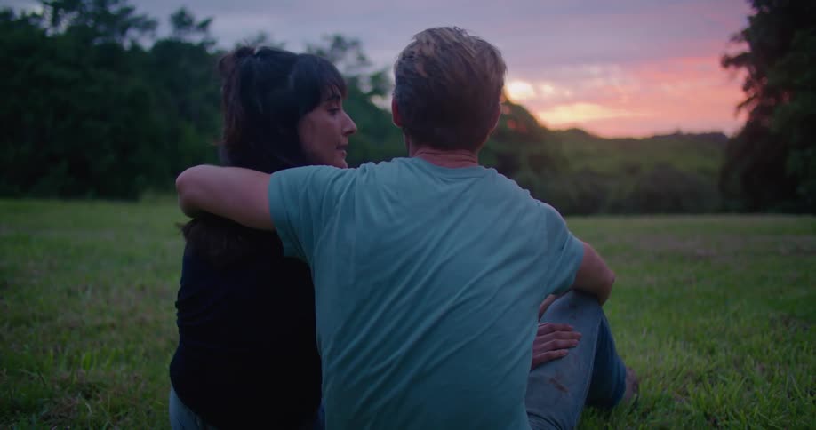 A man and woman kiss while sitting together in a field in the evening