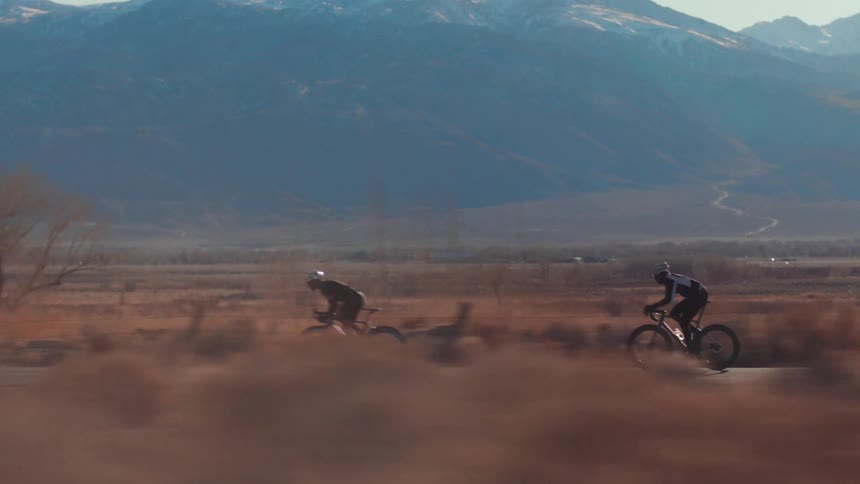 Two cyclists are racing along a road next to mountains topped with snow