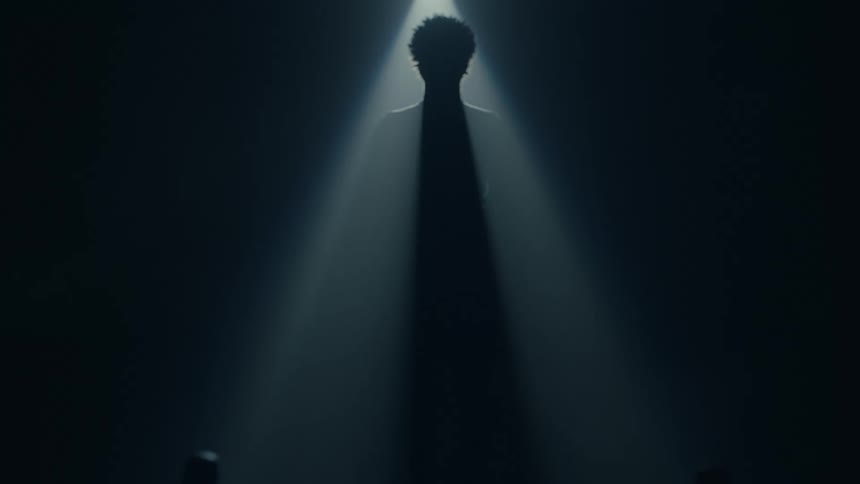Person stands in dark room silhouetted by overhead spotlight near bicycle with headlight on