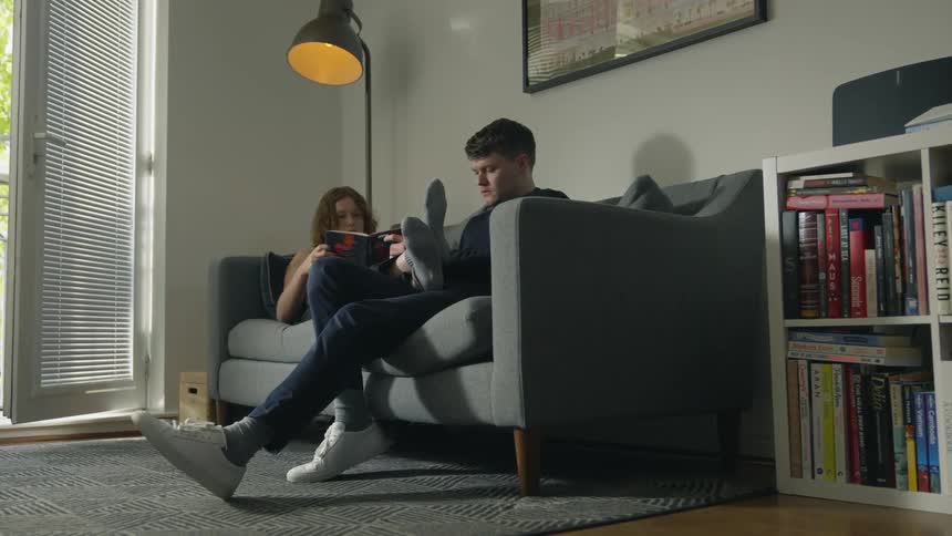 A man sits on a couch and a woman lays beside him reading a book