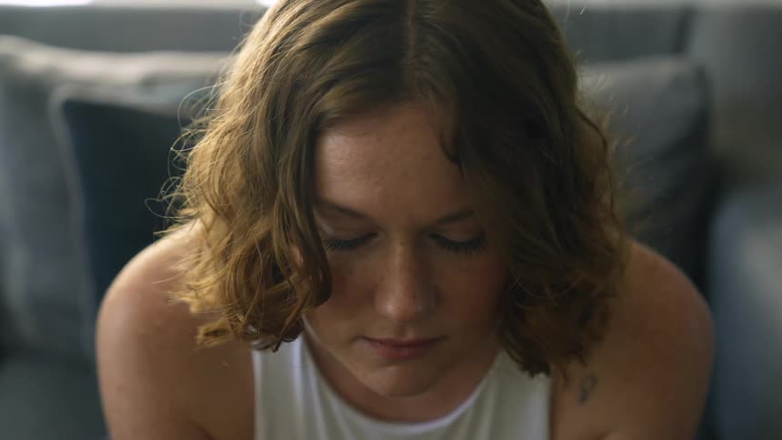 Pensive woman with freckles curly hair and white sleeveless shirt sits on couch in living room at home