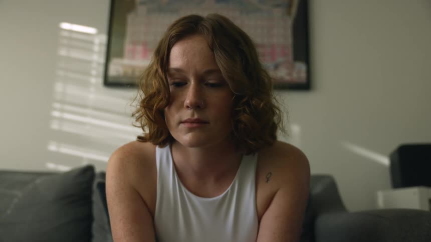 A woman sits on a sofa in the living room of a house while lost in thought