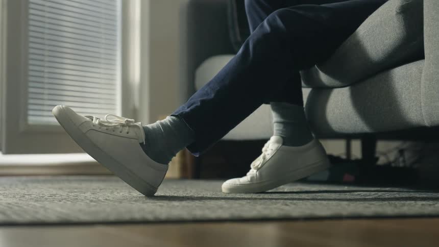 Man with blue pants and white shoes sits on gray coach in living room at home