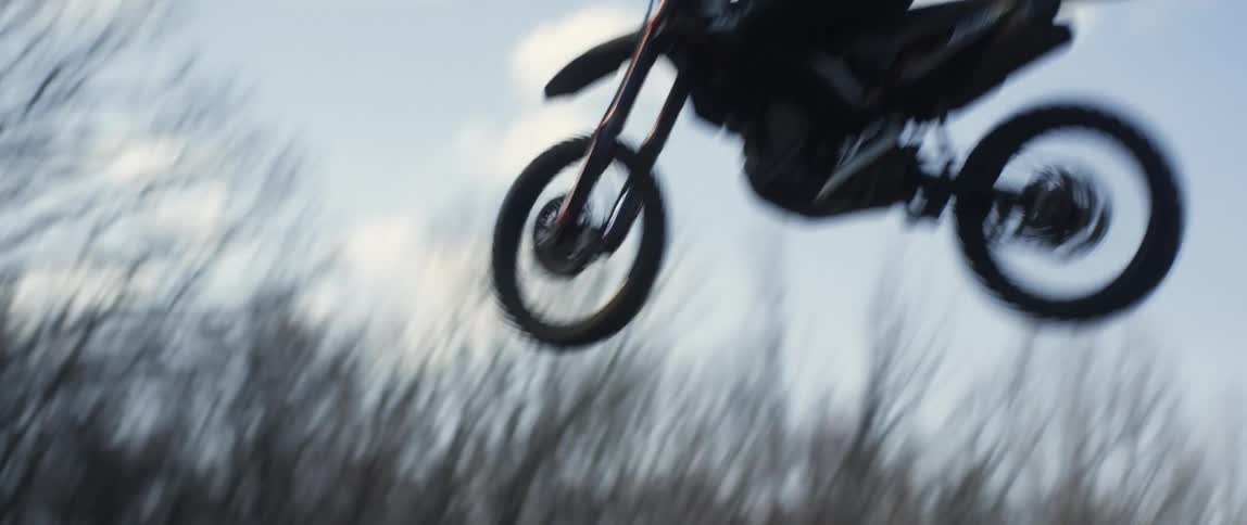 Man rides motorcycle up mound and takes a jump with leafless forest in the background