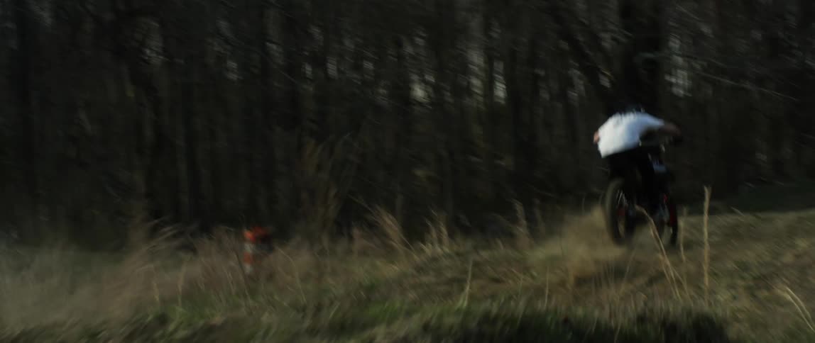 Person jumps motorcycle along hills in field with leafless winter trees in background