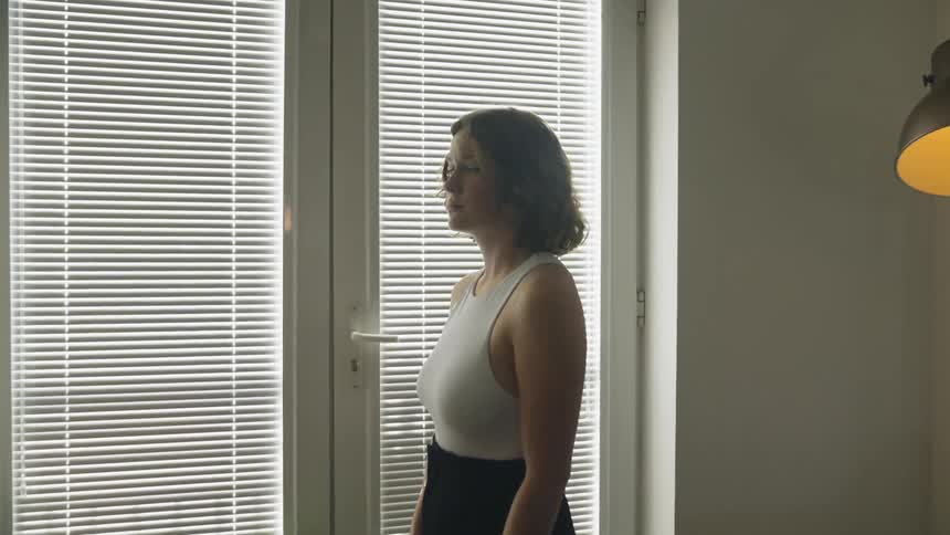 Woman with brown wavy hair white shirt and black skirt stands by windows with blinds in living room at home
