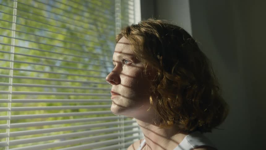 A woman opens and closes her eyes as she stands next to a window