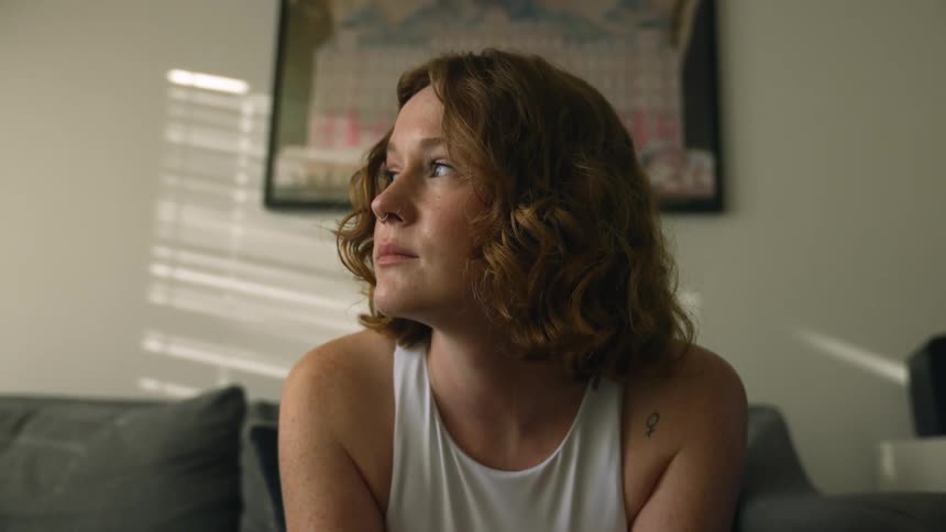 Sad woman with brown wavy hair and white sleeveless shirt sits on couch and looks into space in living room at home