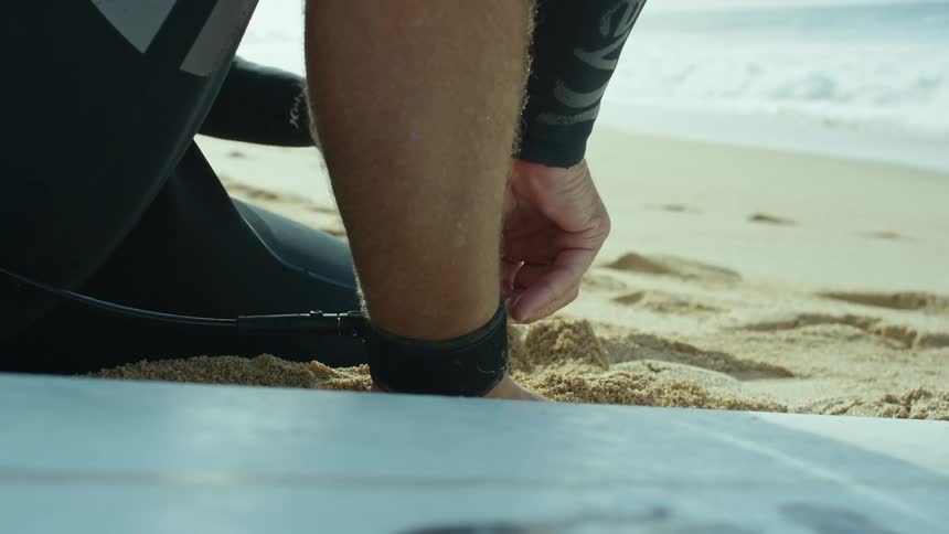 A man sits on the beach and puts a strap on his ankle and then picks up his surfboard