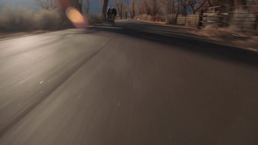 Two male cyclists cycle on country road with fields and distant mountains in the background