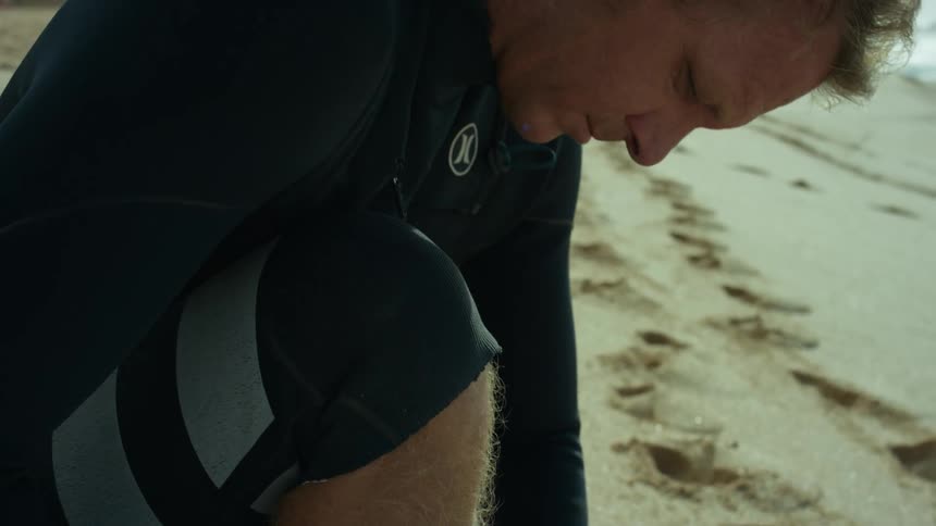 A man straps his surfboard to his ankle while sitting on the beach