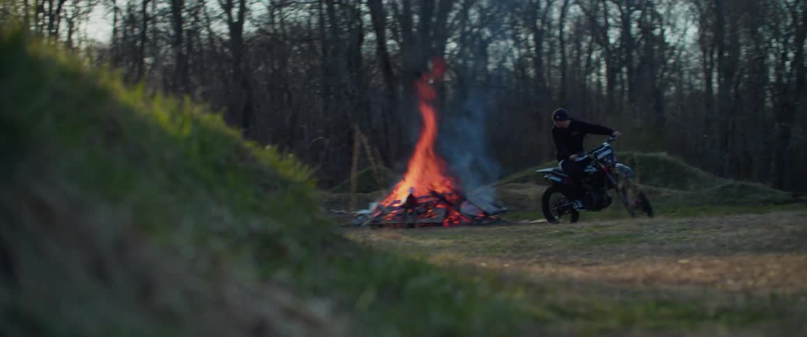Biker rides motorcycle away from smoking bonfire burning in field near leafless trees
