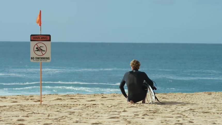 A man sits on the beach near a no swimming sign with a surfboard under his arm