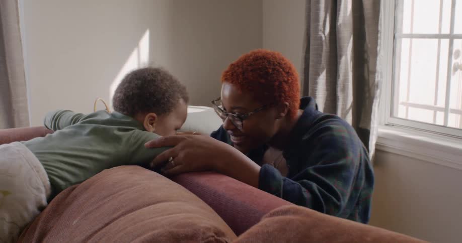 Mother and baby happily play with each other on a couch with sun streaming through windows