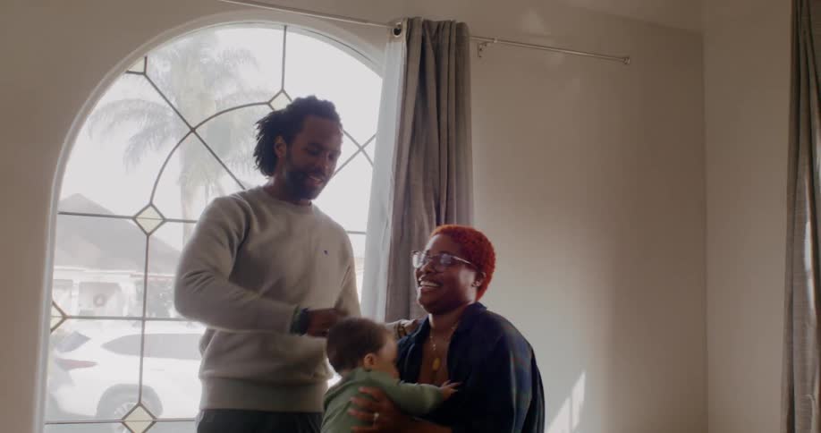 Father with beard gray sweater mother with red hair and black shirt hold toddler baby in room at home