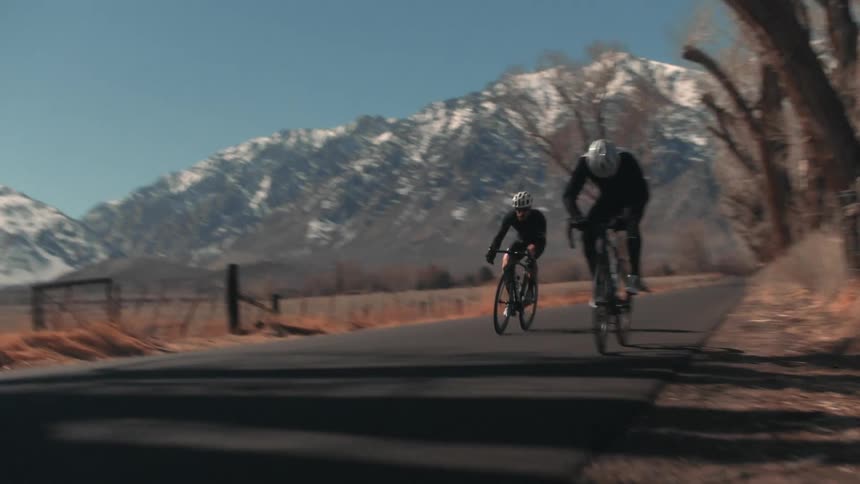 Cyclist jumps on bike and races after another cyclist both dodging car moving along road in the opposite direction