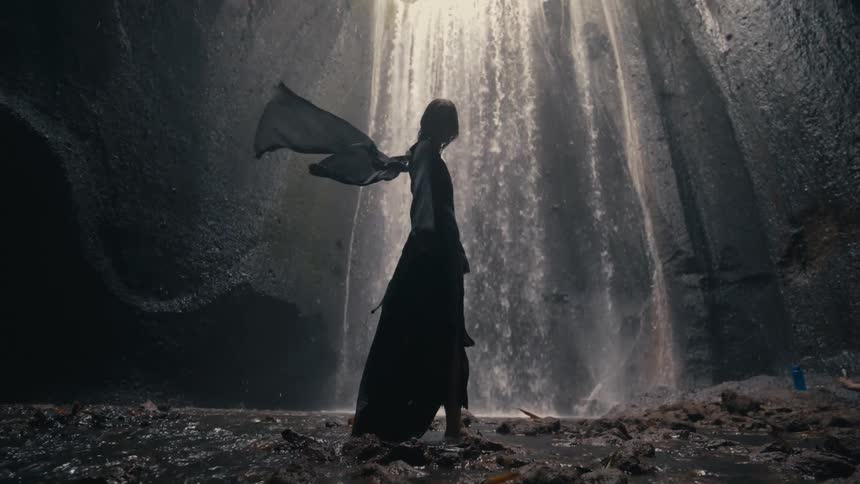 Woman turns and poses near waterfall as another person lies near ground holding object near the wall