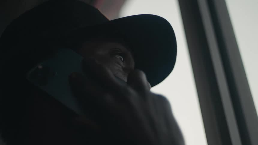 Man with black cap looks out the window talking on smartphone