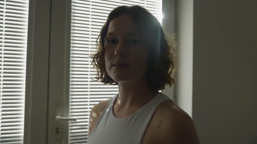 Woman wiht brown curly hair and sleeveless white shirt faces the camera wih eye contact next to windows with blinds