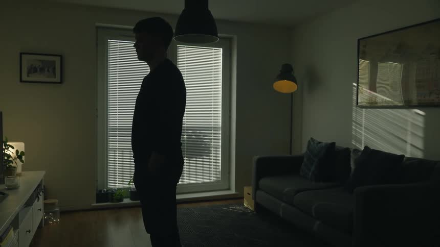 Sad and pensive man standing in living room is silhouetted against the windows with blinds