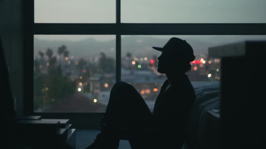 Man sits on floor in dark silhouetted against large window showing hazy town