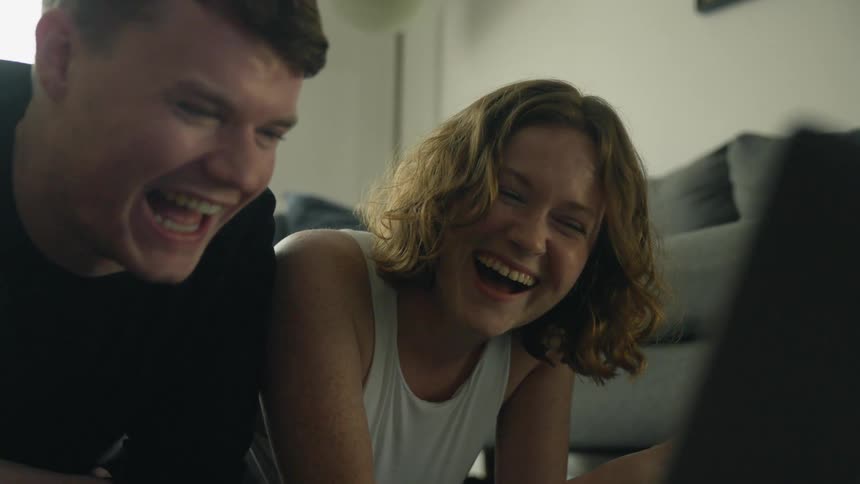 Man and woman lying on their stomachs on the living room floor watching laptop screen start laughing