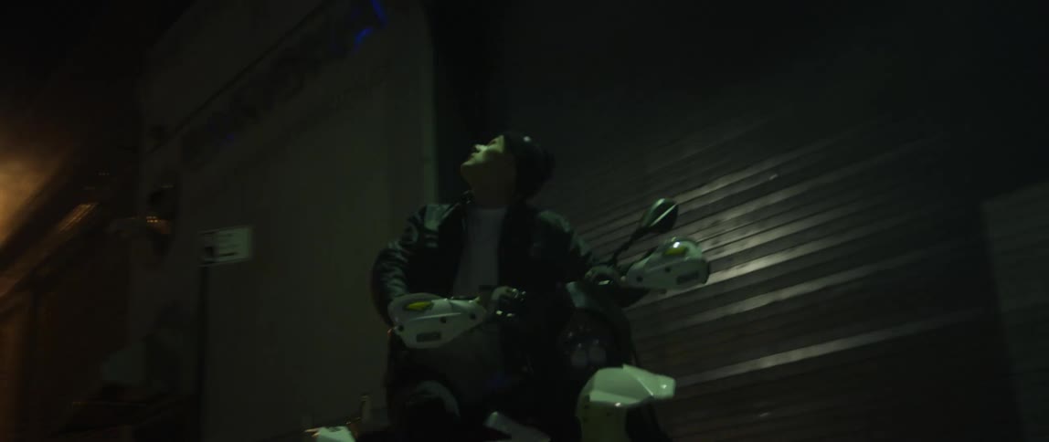 A man stops his scooter in a dark alley and looks up at the buildings