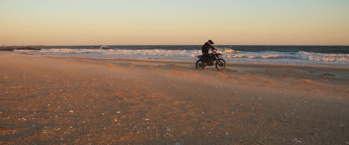 Male biker with helmet black coat and pants rides motorcycle on the beach next to ocean at sunset