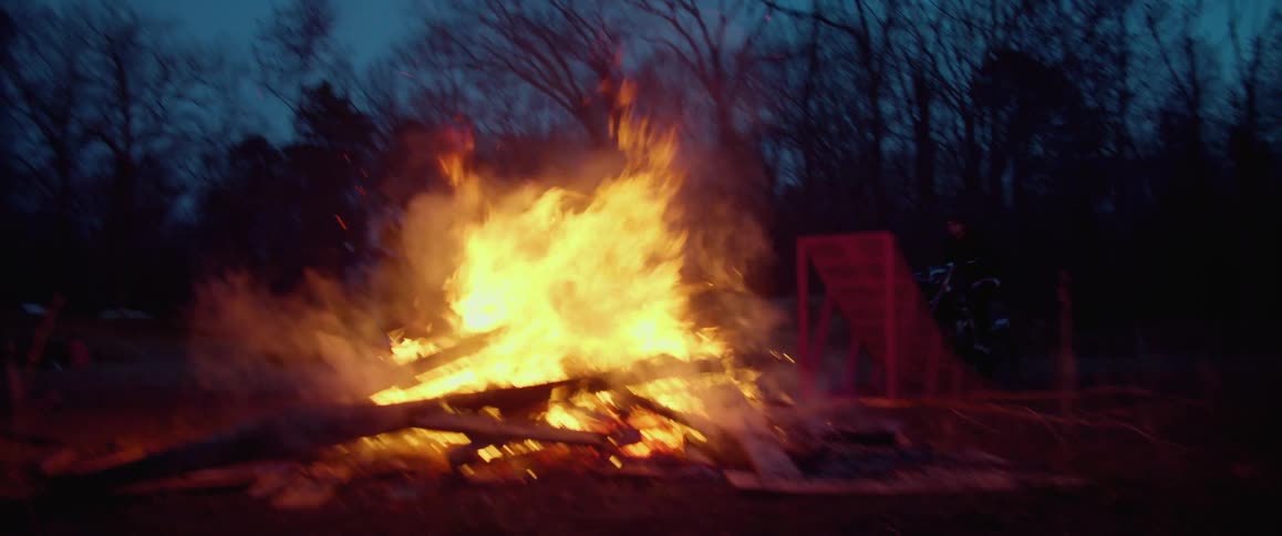 Man rides a motorcycle over a makeshift ramp over a bonfire in the forest at evening