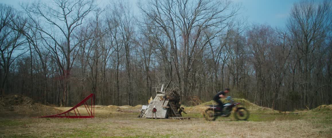 Motorcyclist uses a ramp to jump over pile of junk in field near leafless woods