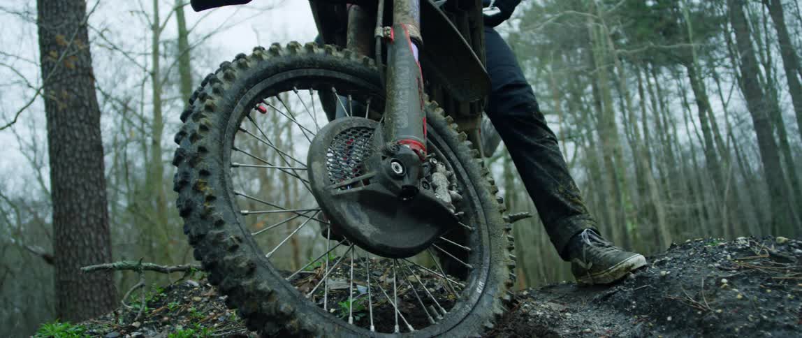 A man stands with a dirt bike on a mound on a dirt trail in a forest on a cloudy day