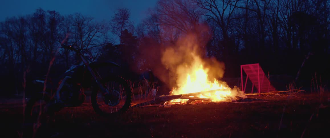 A bonfire burns next to a motorcycle and a ramp with trees in the distance