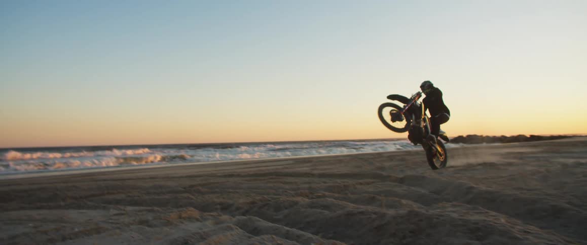 A motorcycle rider does a wheelie across a beach as the waves roll in