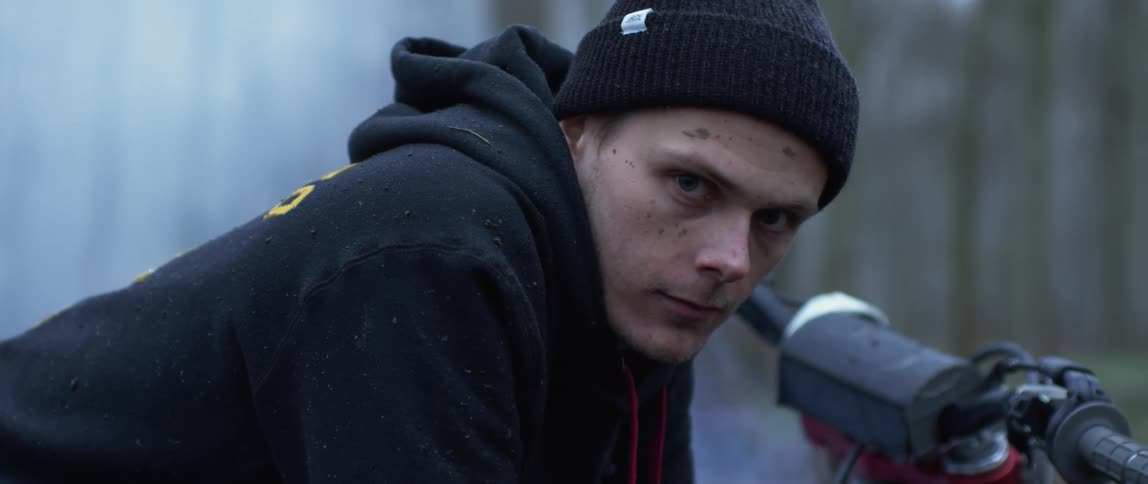 Man wearing hoodie and knit cap leans over bike as smoke rises to obscure trees in the background