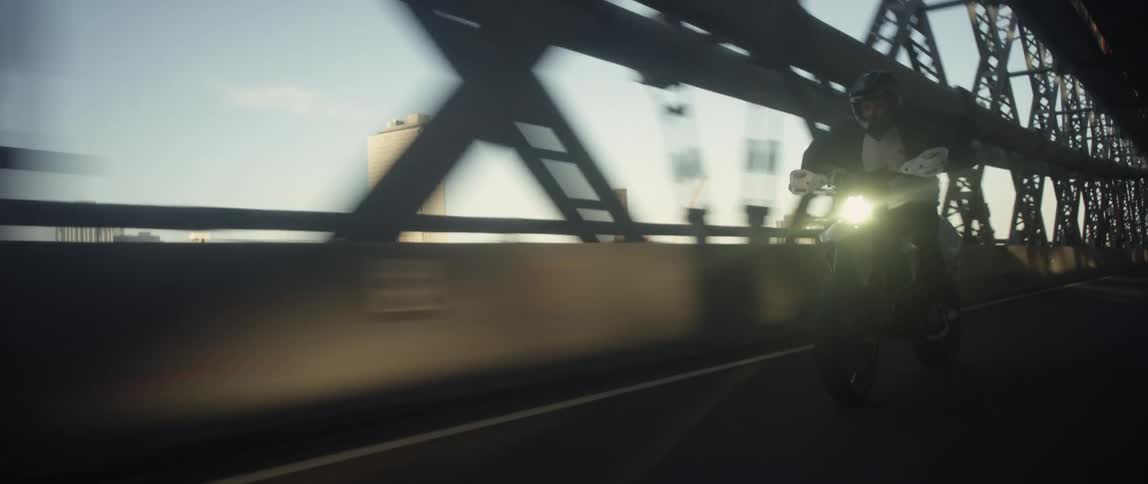 A man rides his motorcycle across a bridge with tall city buildings in the distance