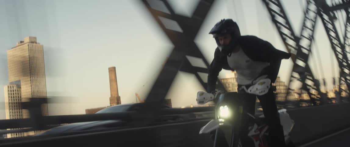 A man is riding a mortorcyle over a bridge in a downtown city