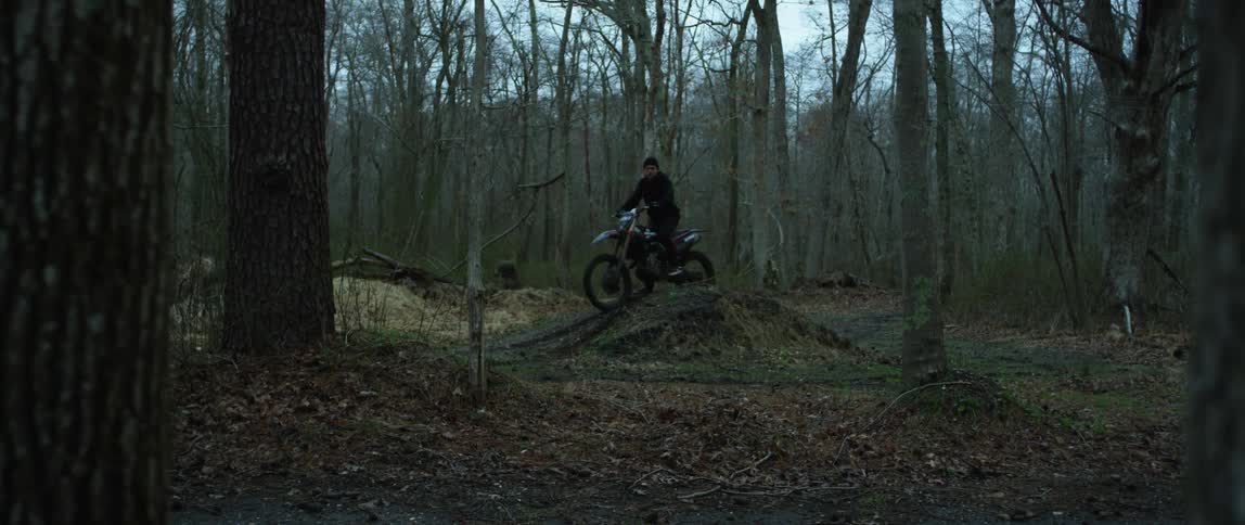 A cyclist riding a motorcycle in the woods rides up a mound and stops at the top